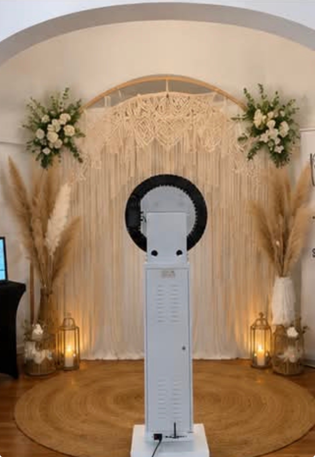 Premium photo booth setup with macramé backdrop, floral arrangements, pampas grass, and gold candle lanterns